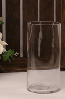 A clear glass cylinder vase placed on a surface with a wooden object in the background.