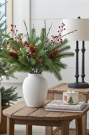 Decorative Christmas arrangement with greenery, red berries, and pinecones in a white vase on a wooden table.