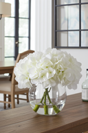 Clear vase with white flowers on a wooden table in a room with large windows.